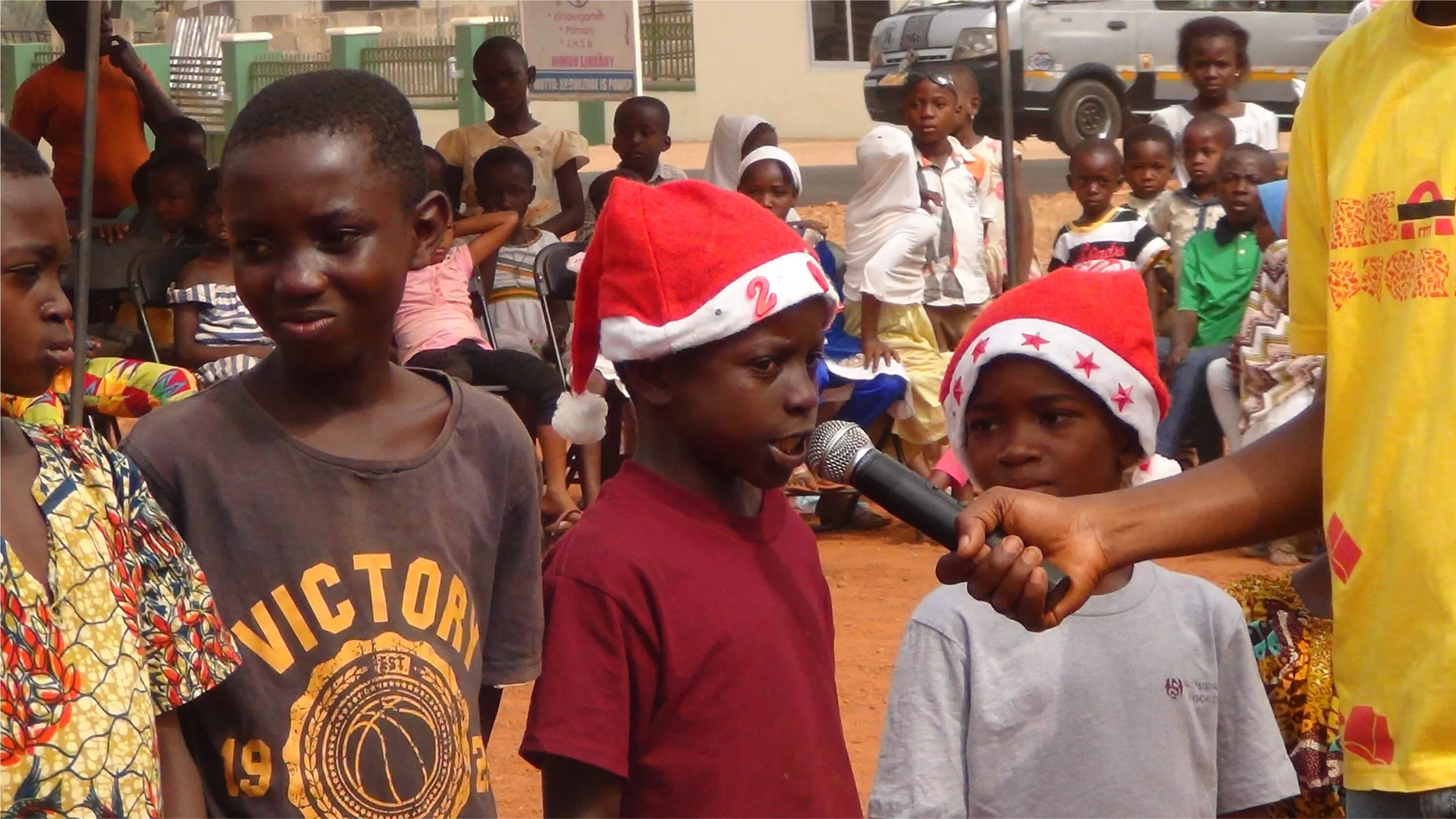 Children in Christmas hats at an outdoor party, one of them speaking into a microphone.