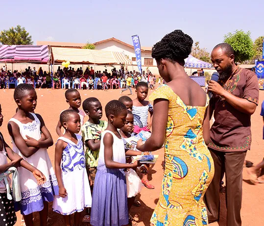 A woman in a yellow print dress handing out gifts to a line of young girls.
