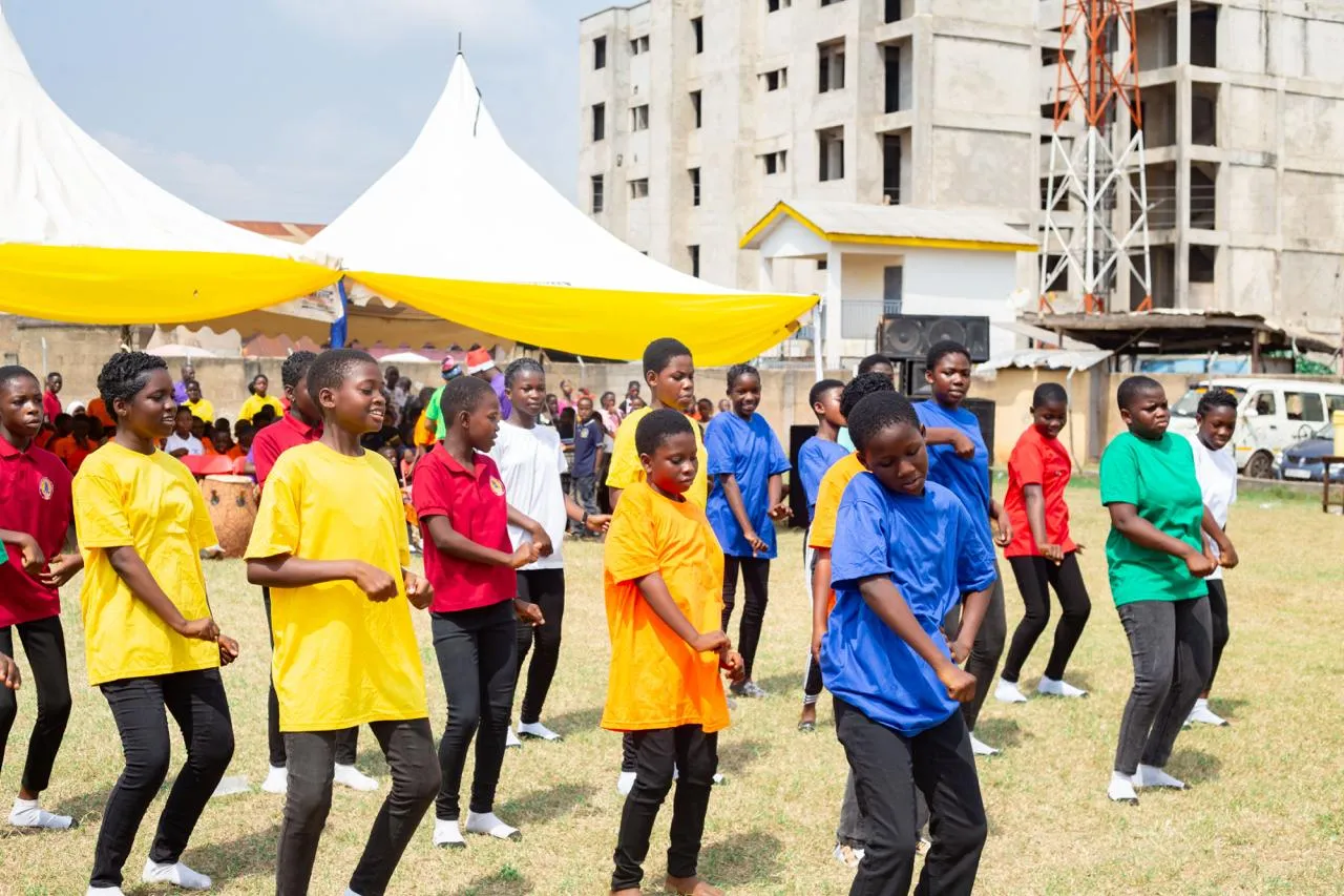 Children in bright yellow, red, blue and green T-shirts dance together on the grass.