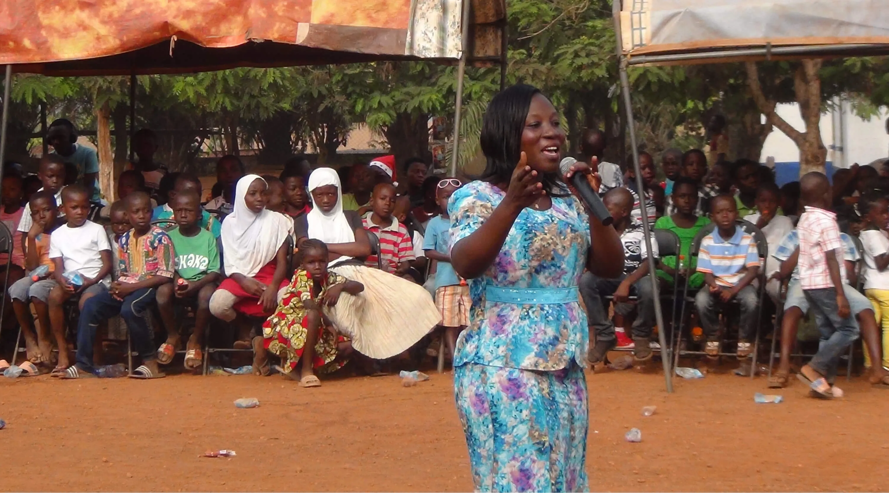 A ministry member speaking into a microphone to children seated under canopies at an outdoor gathering.