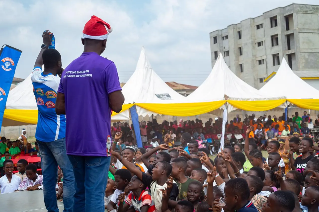A volunteer in a Santa hat faces a large crowd of children cheering under white and yellow tents.