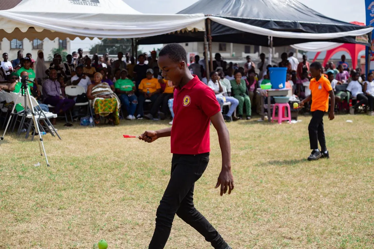 A boy in a red polo shirt carries a spoon in a race while families watch from under the tents.